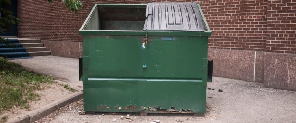 Green dumpster in an urban setting surrounded by brick walls and concrete pathways during daylight hours